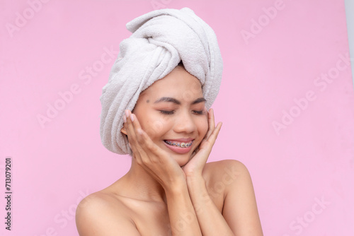 Radiant young Asian woman with head wrapped in a towel and dental braces, exuding happiness and self-care on a pink background.