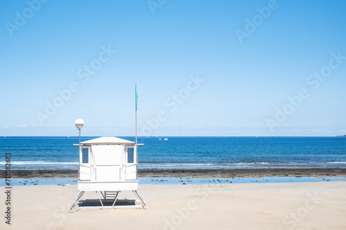 Fototapeta Naklejka Na Ścianę i Meble -  white lifeguard tower on a deserted beach. The clear sky and calm sea create a serene backdrop, with a green flag indicating safe swimming conditions.