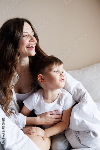 Portrait of smiling woman hugging cute toddler son under blanket on bed in morning