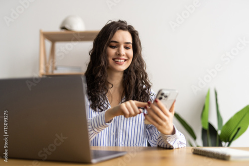 Young smiling business woman sitting at table in coffee shop and uses smartphone.