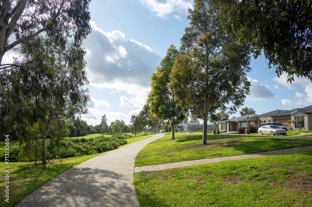 Obraz premium A pedestrian walkway in a public park with some residential houses in the distance. A suburban park with a walking trail in an Australian residential neighbourhood. Beautiful living environment. 