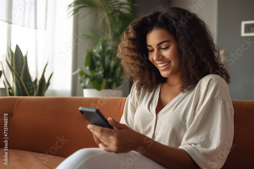 Happy young Latin woman sitting on sofa holding mobile phone using cellphone technology doing ecommerce shopping, buying online, texting messages.