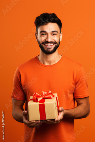 Young smiling happy man wearing t-shirt casual clothes hold present box with gift ribbon bow looking aside on area isolated on orange red color background studio portrait. © AI_images