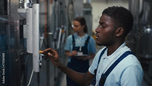 Tilt side footage of African American engineer in polo shirt and jumpsuit adjusting mechanism operation using sensor monitor in service room at plant photo