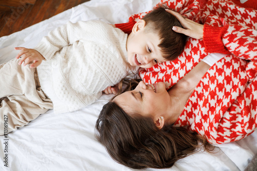 Cheerful mother and toddler son in knitwear talking while relaxing on bed