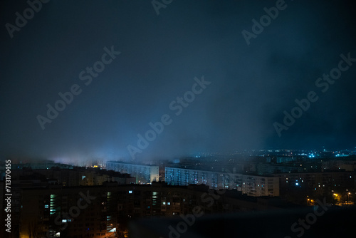 The twinkling lights of a bustling city are captured at night  reflecting off distant water and contrasting with the dark  silhouetted buildings.