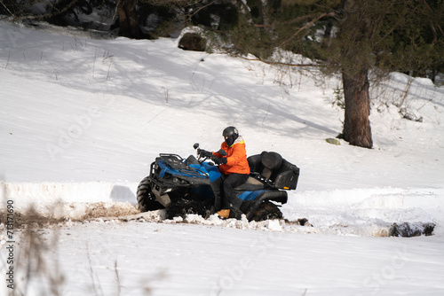 A man, equipped with cold-weather attire, confidently handles a red quad bike along a snowy path, surrounded by a wintry forest landscape.