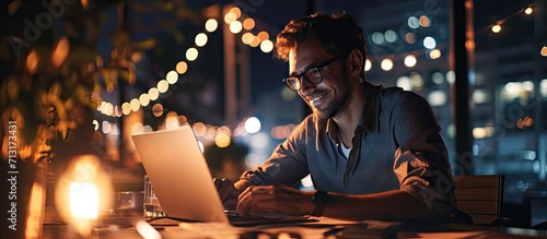 Joyful young businessman sitting at desk looking at computer screen talking with business partners make informal video call worker have online meeting with colleagues working late at dark offic