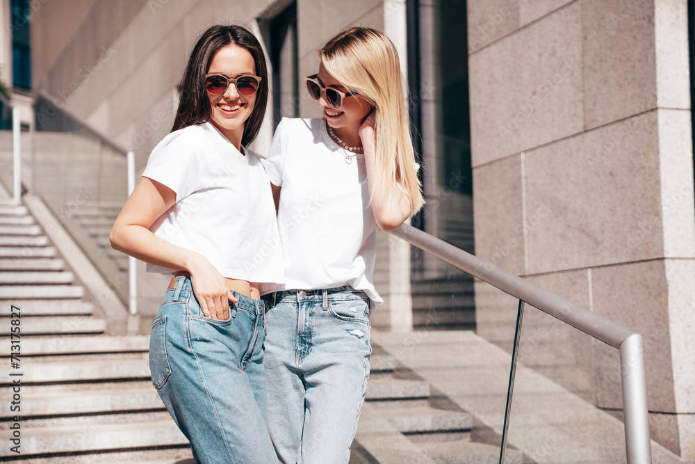 Two young beautiful smiling hipster female in trendy summer white t-shirt and jeans clothes. Carefree women posing in street. Positive models having fun. Cheerful and happy. In sunglasses
