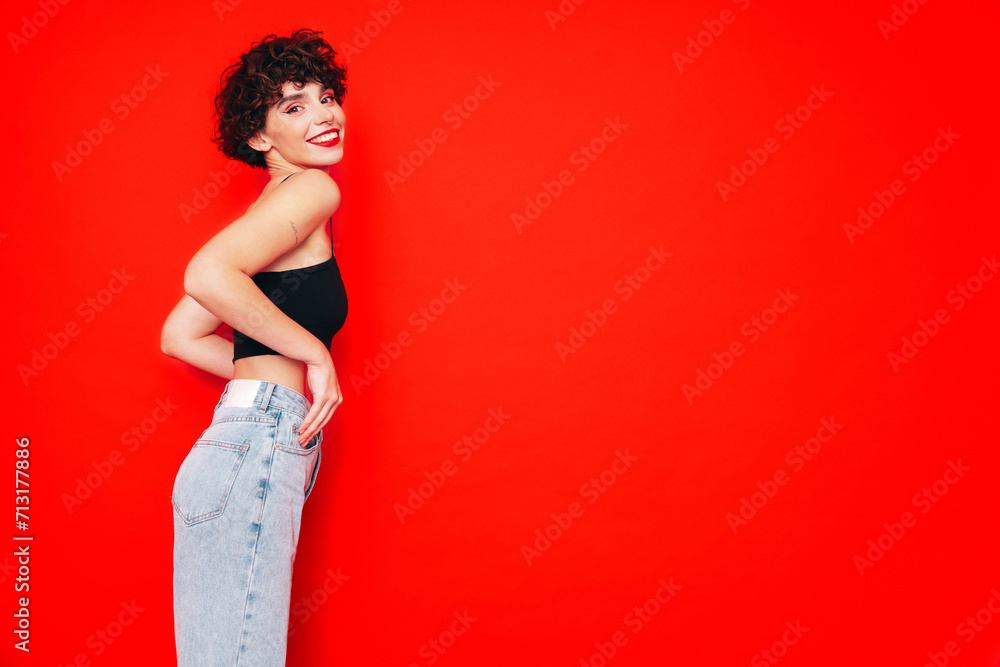 Young beautiful smiling female in trendy summer black tank top. Carefree woman posing near red wall in studio with curly hairstyle. Positive model having fun. Cheerful and happy Young beautiful smiling female in trendy summer black tank top. Carefree woman posing near red wall in studio with curly hairstyle. Positive model having fun. Cheerful and happy