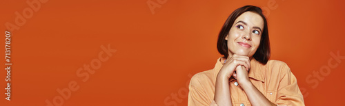 hopeful young woman with short hair smiling and looking away on orange background, banner