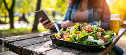 Close up woman using meal tracker app on phone while eating salad at picnic table in the park on a break Healthy balanced diet lunch box Healthy diet plan for weight loss Selective focus photo