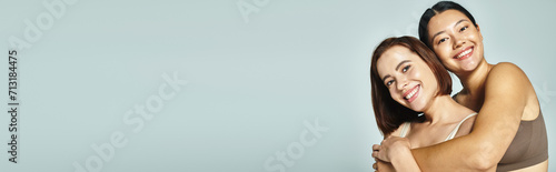 joyful asian woman in underwear embracing her young friend on grey background, companionship banner