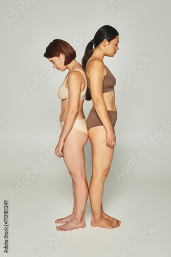 upset multicultural women in underwear standing back to back on grey backdrop, body shaming
