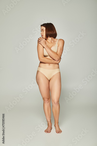 full length of shy woman in beige underwear looking over shoulder on grey background, vulnerability