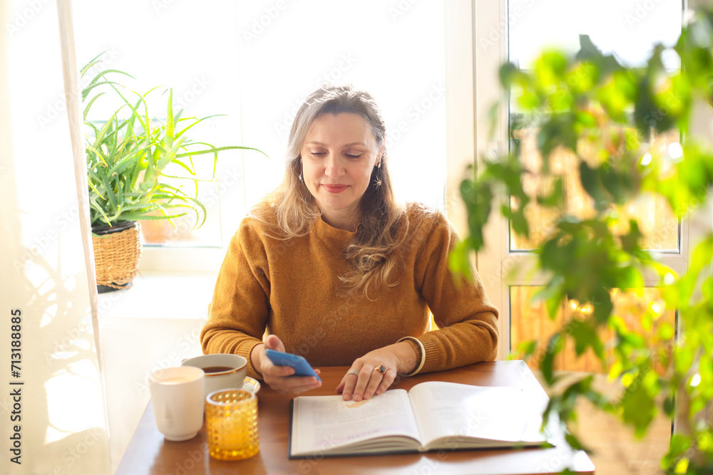 Smiling middle-aged woman surfing internet on smartphone screen while reading book at home Smiling middle-aged woman surfing internet on smartphone screen while reading book at home