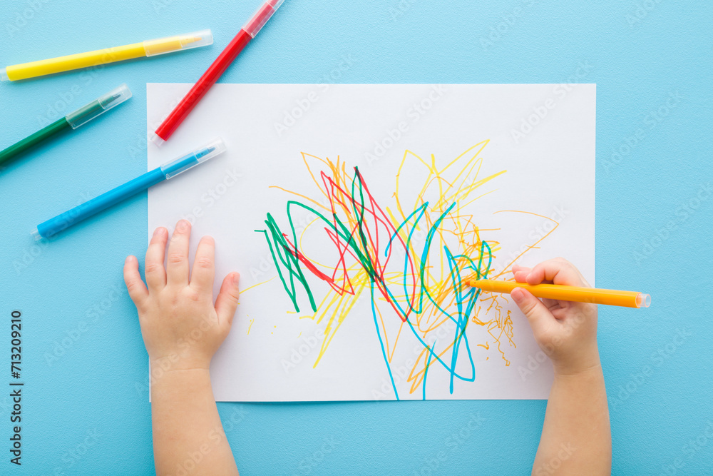 Baby hand holding marker and drawing colorful scratches lines on white paper. Blue table background. Pastel color. Closeup. Toddler development. Learning painting. Point of view shot. Top down view. - obrazy, fototapety, plakaty 