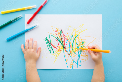 Baby hand holding marker and drawing colorful scratches lines on white paper. Blue table background. Pastel color. Closeup. Toddler development. Learning painting. Point of view shot. Top down view. photo
