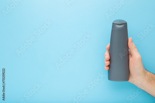 Young adult man hand holding gray plastic shampoo or shower gel bottle on blue background. Pastel color. Care about clean body skin. Male beauty product. Closeup. Empty place for text. Top down view.