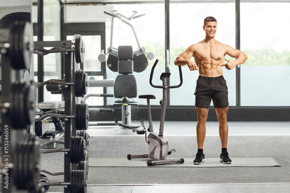 Coach posing shirtless and leaning on an exercise bike at a gym