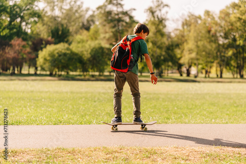 Back view shot of a cool teen boy learning to ride his skateboard in sunlight in a park.