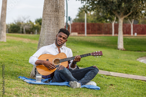 An African American man singing and enjoying nature while playing a guitar and sitting on the grass in a local park.