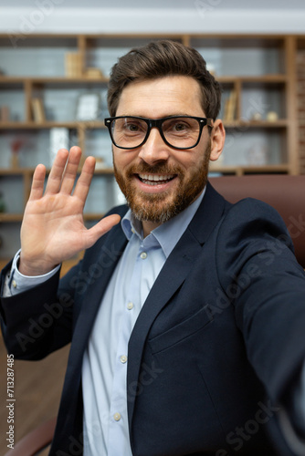 Mature businessman in glasses and suit smiling and waving at the camera, giving a friendly greeting from his cozy office.