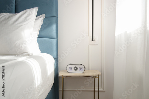 Blue king size bed with crisp white bedding and digital alarm clock on the nightstand under beautiful morning sunlights	 photo