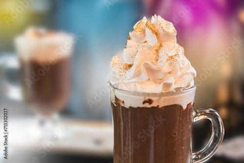 Hot chocolate with whipped cream and edible gold glitter in a transparent cup. Blurred colorful background.