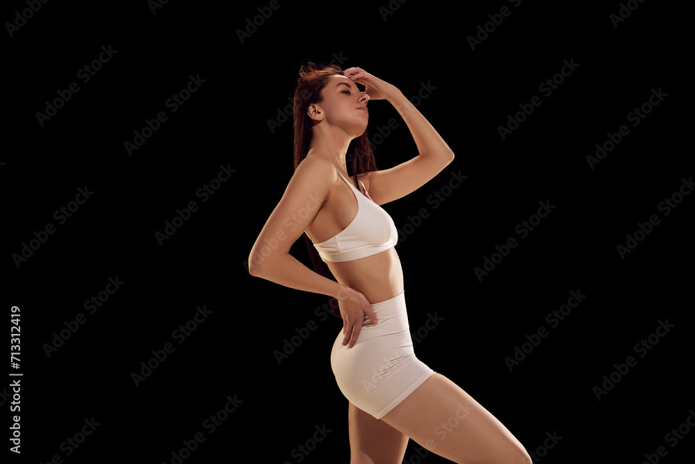 Fototapeta premium Slim, beautiful female model posing against black studio background. Woman wearing in sporty clothes, shorts and top. Concept of female health, fitness, sport, active lifestyle, dieting.