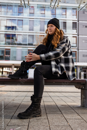 Woman in her mid-thirties with long red hair wearing a cap and black clothing in an urban location.