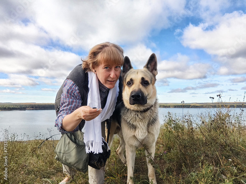 Adult girl with shepherd dog taking selfie near water of river or lake. Middle aged woman and big pet on nature. Friendship, love, fun, hugs © keleny
