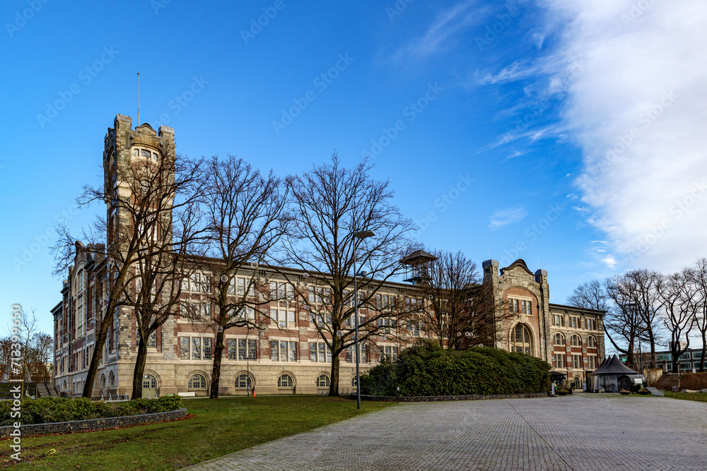 Empty esplanade with old main building of former Waterschei mine in background against blue sky, bare trees, Thor Park - Hoge Kempen National Park, sunny autumn day in Genk, Belgium