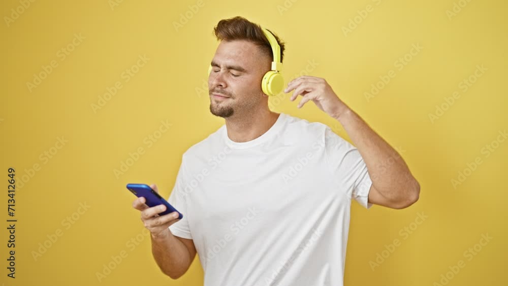 A smiling young man with a beard wearing headphones and holding a smartphone against a yellow background. A smiling young man with a beard wearing headphones and holding a smartphone against a yellow background.