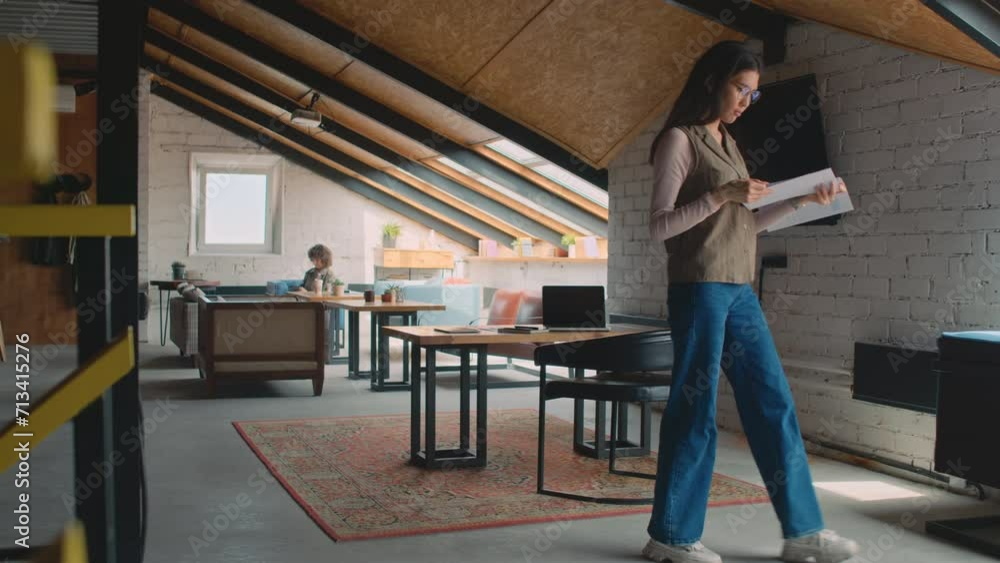 Medium full shot of Asian woman standing in loft office, reading report, African American man with laptop walking up and showing something to her, then leaving and sitting down at tableMedium full sho Medium full shot of Asian woman standing in loft office, reading report, African American man with laptop walking up and showing something to her, then leaving and sitting down at tableMedium full sho