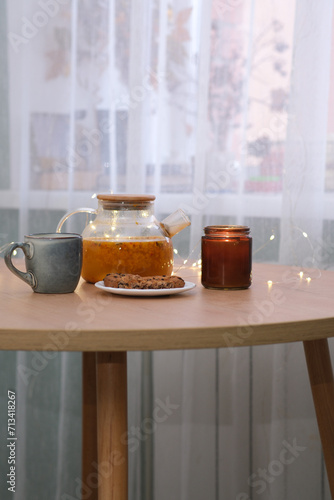 Home kitchen interior- glass hot teapot with sea buckthorn tea   plate with biscuits  burning candle on wooden table near the window.