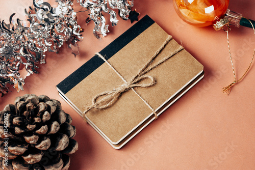 Christmas gift. Kraft paper notebook, silver tinsel on peach pastel table. New Year 2024 present, pine cone. Festive decor. Holidays congratulations.