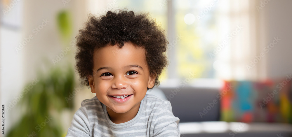 Fototapeta premium Portrait of a happy cute African American kid with afro fluffy hair looking at the camera. Black beautiful happy healthy child. A child sits in a children's room with toys, in a private kindergarten.