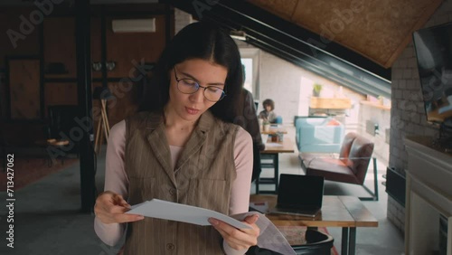Medium shot of young Asian woman in glasses standing in coworking office, holding document printout or presentation, reading then sighing, black man with laptop entering and sitting down at tableMediu photo