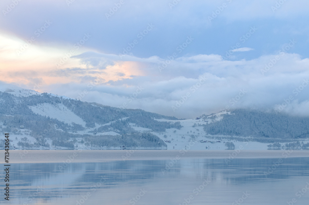 A tranquil scene captures the serene beauty of a snow-covered landscape and a calm fjord under a soft winter sunset with fog lifting off the waters surface.
