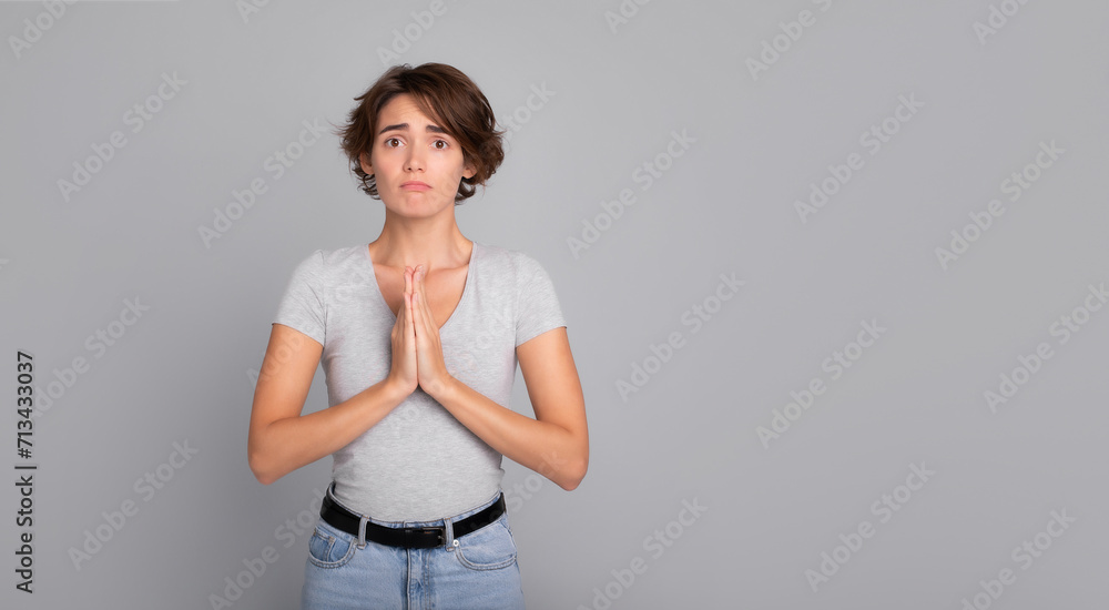 Attractive young woman looks in camera and stands in meditative pose, enjoys peaceful atmosphere, hands in praying gesture and asking help, isolated over gray background, peace in soul Attractive young woman looks in camera and stands in meditative pose, enjoys peaceful atmosphere, hands in praying gesture and asking help, isolated over gray background, peace in soul