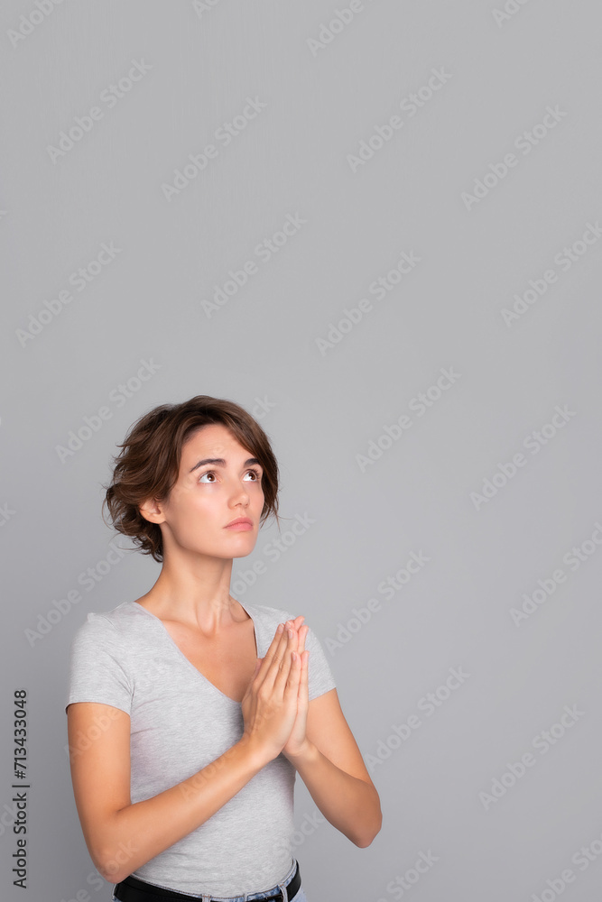 Vertical photo of cute young woman stands in meditative pose, enjoys peaceful atmosphere, hands in praying gesture and asking help, isolated over gray background, peace in soul Vertical photo of cute young woman stands in meditative pose, enjoys peaceful atmosphere, hands in praying gesture and asking help, isolated over gray background, peace in soul