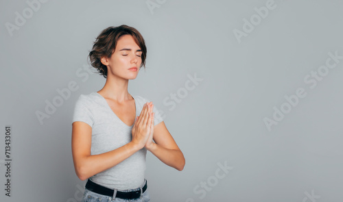 Peaceful young girl with closed eyes stands in meditative pose, enjoys peaceful atmosphere, hands in praying gesture and asking help, isolated over gray background, peace in soul