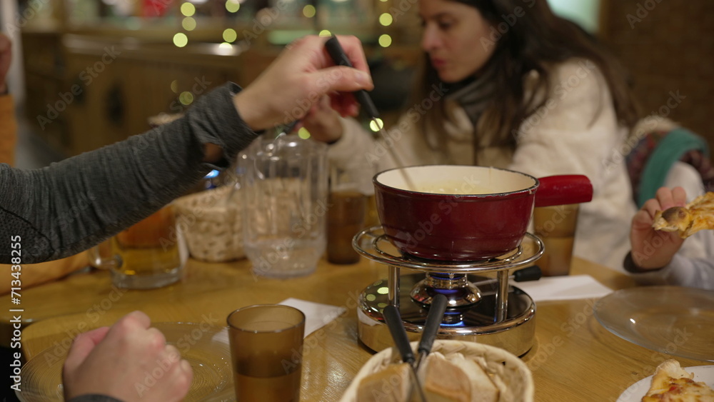 People eating cheese fondue at restaurant, closeup hand dips piece of bread into melted cheese at diner, red pot on top of heater