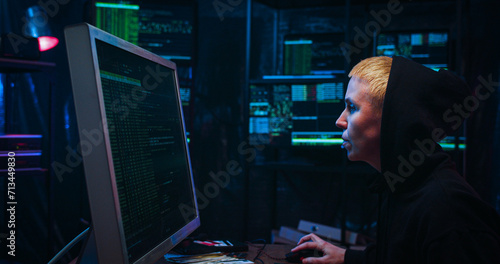 Side view on Caucasian woman with short blond hair and in hood working in cyber security center and analyzing data. Female hacker typing on keyboard at big computer in dark monitoring room.