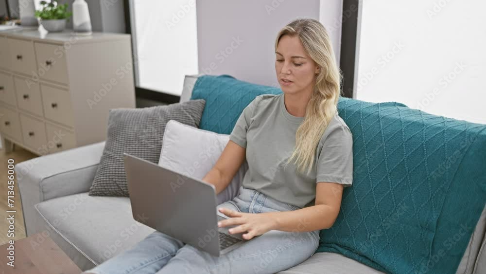 A relaxed blonde woman using a laptop on a couch in a modern living room setting. A relaxed blonde woman using a laptop on a couch in a modern living room setting.