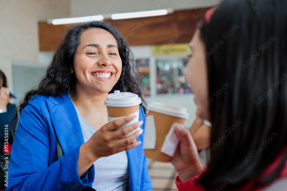 Two young smiling latin women commuters waiting for public train transportation in the station while drinking hot coffee Two young smiling latin women commuters waiting for public train transportation in the station while drinking hot coffee