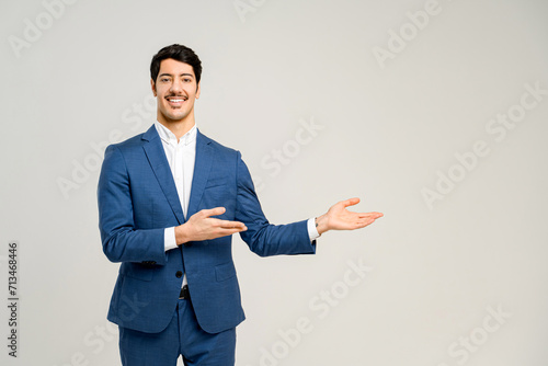 Young male business professional in a stylish blue suit pointing aside, his open hand suggesting a presentation or a welcoming introduction, set against a plain background photo