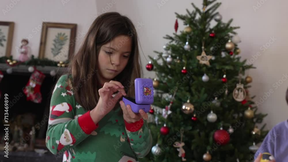 Little girl absorbed by Christmas present standing in living room while family opens gifts around her, holiday Christmas tree in background Little girl absorbed by Christmas present standing in living room while family opens gifts around her, holiday Christmas tree in background