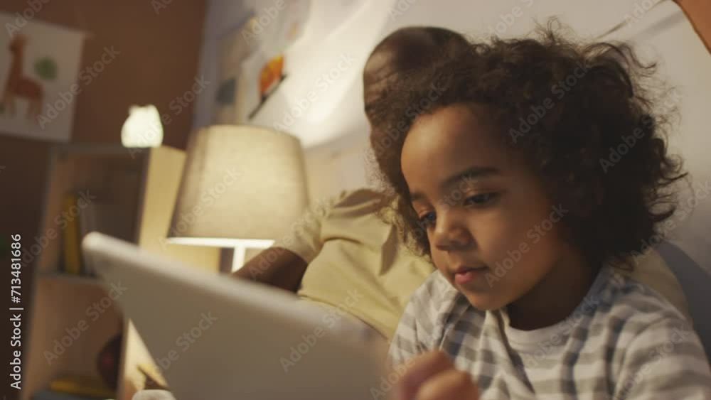 Side shot of African American little curly boy in pajamas using tablet sitting with dad in bed before sleep in cozy bedroomSide shot of African American little curly boy in pajamas using tablet sittin Side shot of African American little curly boy in pajamas using tablet sitting with dad in bed before sleep in cozy bedroomSide shot of African American little curly boy in pajamas using tablet sittin
